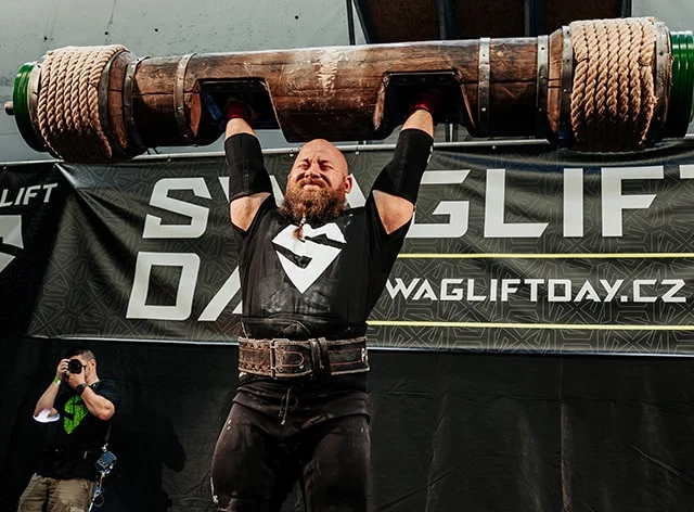 A competitor lifts a&nbsp;massive wooden log overhead in front of a&nbsp;SWAGLIFT DAY banner